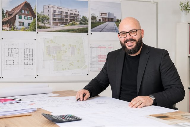 Easyhomes-Projektentwickler am Schreibtisch, im Hintergrund die Visualisierungen von Bauernhaus, Mehrfamilienhaus und Einfamilienhaus